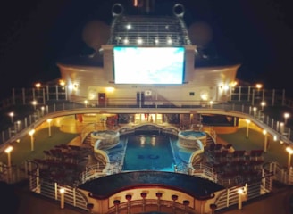 A luxury cruise ship deck featuring a pool surrounded by lounge chairs and warm ambient lighting. A large screen overlooks the area, showing an image against a dark night sky.
